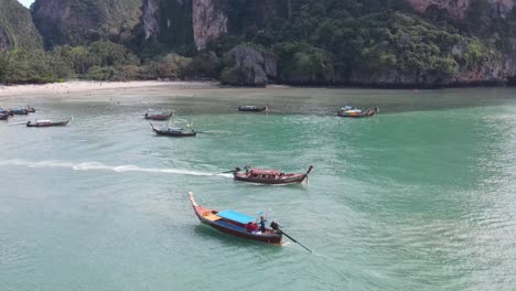 Aerial-View-Of-Longtail-Boat-Leaving-Beach-With-Pan-Reveal-Of-Limestone-Cliffs-In-Background-At-Railay,-Thailand