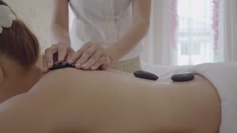 Professionelle-Hot-Stone-Massage,-Therapeut-Massiert-Personen-Mit-Steinen-Zurück,-Während-Sie-Auf-Dem-Massagetisch-Liegen,-Wohlbefinden-Und-Gesundheitskonzept