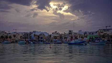 Blue-white-boats-anchored-in-port-town-as-sun-sets-over-Marsaxlokk,-Malta,-timelapse