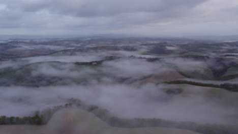 Rolling-hills-of-Tuscany-covered-in-mist-before-sunrise,-aerial