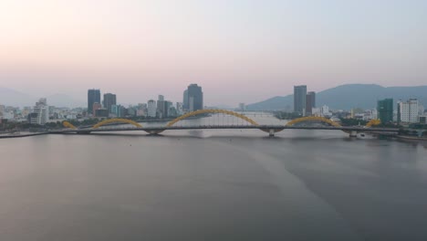 Colorful-wide-aerial-of-iconic-Dragon-Bridge-Cau-Rong,-traffic-and-city-skyline-during-sunset-in-Danang,-Vietnam