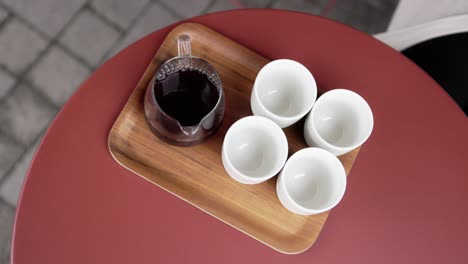 Glass-carafe-and-four-white-cups-are-arranged-on-wooden-tray-on-red-table