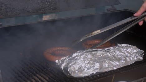 Tilt-down-to-tongs-moving-BBQ-sausage-around-on-smoking-BBQ-pit