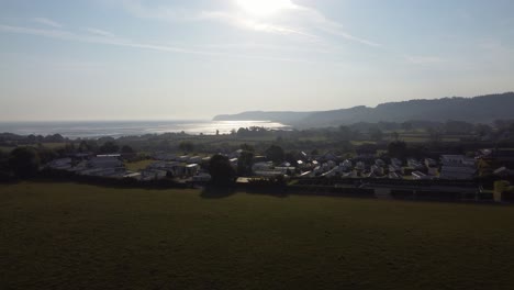 Rising-aerial-view-Welsh-caravan-park-at-sunrise-overlooking-shimmering-coastal-bay-and-rolling-countryside-hills