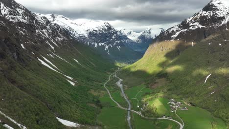 Hjelledalen-Valley-with-Strynevegen-leading-to-Strynefjell-mountain---Springtime-aerial-with-snow-capped-mountains-and-spots-of-sun-in-green-hillsides