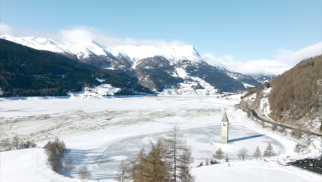 Aerial-Flying-Over-Church-Bell-Tower-With-View-Of-Frozen-Lake-Reschen-And-Submerged-Tower-On-Clear-Sunny-Day