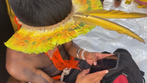 Close-up-shot-of-a-young-amazonian-man-wearing-traditional-colorful-feather-hat-holding-his-smartphone
