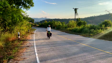A-lone-female-cyclist-rides-along-an-empty-country-road-near-Paknai's-Fisherman-Village-in-the-Na-Muen-District,-Nan,-Thailand