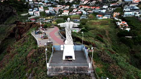 Establishing-aerial-view-Cristo-Rei-statue-on-Madeira-island-hillside-overlooking-Garajau-ocean-coastline