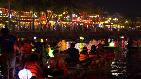 Traditional-Vietnamese-row-boats-on-river-with-illuminated-Hoi-An-city-in-distance