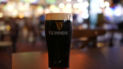 Una-Pinta-De-Cerveza-Guinness-Stout-Ale-En-Una-Mesa-En-Un-Pub-Irlandés-El-Día-De-San-Patricio-En-Irlanda-Con-Clientes-Caminando