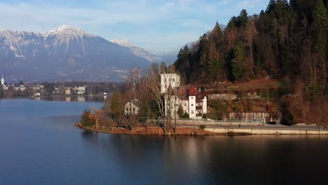 Aerial-Lakeside-View-Of-Adora-Luxury-Hotel-With-Pan-Left-Reveal-To-Bled-Castle-And-Snow-Capped-Karawanks-Mountain-Range-In-Background