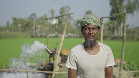 Ein-Armer-Bangladeschischer-Bauer-Wirft-Einen-Blick-In-Die-Kamera,-Während-Im-Hintergrund-Eine-Wasserpumpe-Läuft,-Die-Wasser-In-Das-Erntefeld-Spült