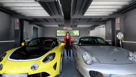 in-a-garage,-a-dark-haired-woman-dressed-in-black-and-red-walks-between-two-luxury-cars-of-well-known-brands,-while-talking-in-front-of-the-camera