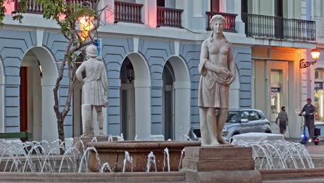 Street-view-of-the-Plaza-de-Armas-fountain-with-statues-as-pedestrians,-cars-and-a-scooter-transit-in-the-background