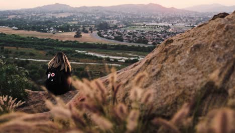 girl-sitting-on-a-cliff-edge-with-her-hair-blowing-in-the-wind-at-sunset-with-foxtail-grass-in-the-foreground