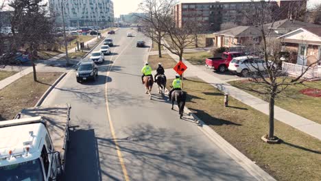 Aerial-tracking-shot-of-Canadian-police-horsemen-patrolling-down-a-busy-road-in-Toronto,-bright-daylight