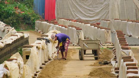 Brick-field-In-Bangladesh,-lonely-worker-with-shovel-loading-handcart-with-sand
