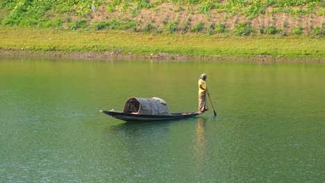 Pescador-Bengalí-Parado-En-El-Borde-De-Su-Barco-Rural-Buscando-Peces-En-Un-Río-Que-Fluye-Lentamente-Con-Hierba-Viva-En-El-Fondo,-Bangladesh
