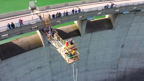 Aerial-drone-view-of-young-people-bungee-jumping-off-a-50m-high-dam-platform-while-viewers-standing-on-dam-bridge-watching-action-happen