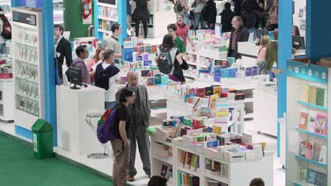 Brightly-lit-book-display-shelfs-in-center-of-bookfair,-people-walk-through-searching