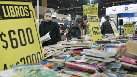 Two-women-framed-between-discount-table-signs-at-book-fair-with-haphazard-stacked-books-on-table