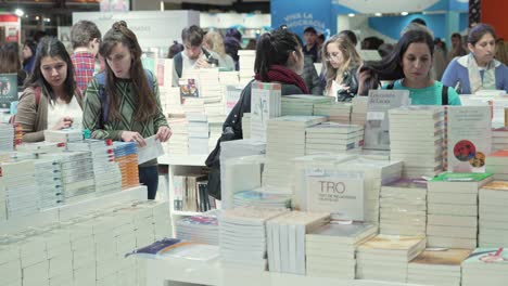 Women-and-men-walk-through-piles-of-books-piled-high-in-convention-center-expo