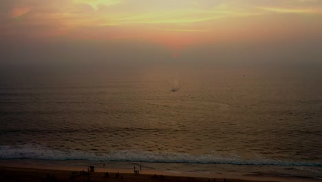 Aerial-camera-moving-zoom-in-into-Rescue-lifeguard-in-the-ocean-in-front-of-a-big-wave-searching-for-surfers,-Sunset-of-dramatically-orange-sky