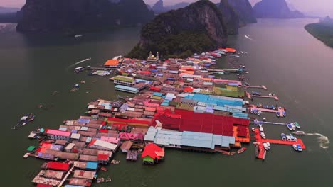 Panyee-floating-village-aerial-view-in-which-the-carrier-aerial-drone-camera-is-moving-behind,-showing-the-colorful-village-and-many-boats-ferrying-tourists-around