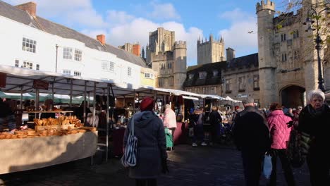 People-shopping-and-wandering-around-vendor-stalls-at-busy-market-place-in-city-of-Wells,-Somerset-England