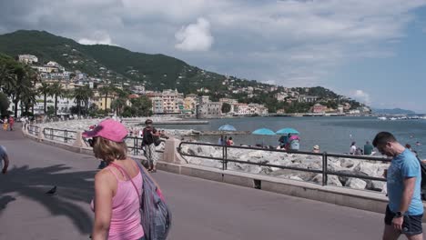 Menschen-Gehen-Auf-Der-Promenade-In-Rapallo,-Italien,-Während-Andere-Am-Strand-Ein-Sonnenbad-Nehmen