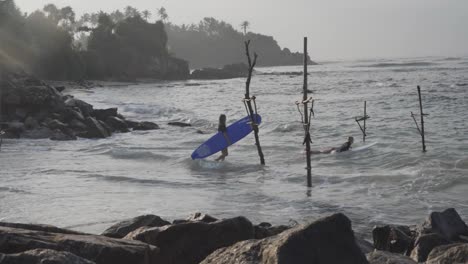 Surfers-Walking-Towards-The-Ocean-And-Surfing-At-The-Weligama-Coast-With-Wooden-Stilts-In-Matara,-Sri-Lanka