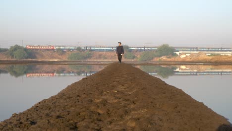 Guy-Walking-On-The-Muddy-Walkway-Between-Ponds-In-Mumbai,-India-With-Local-Train-Running-In-The-Background-On-A-Sunny-Morning---long-shot