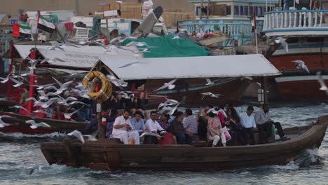 Tourists-Riding-The-Famous-Abra-Ferry-Boats-Sailing-On-The-Dubai-Creek-With-Seagulls-Flying-Around-In-Dubai,-UAE