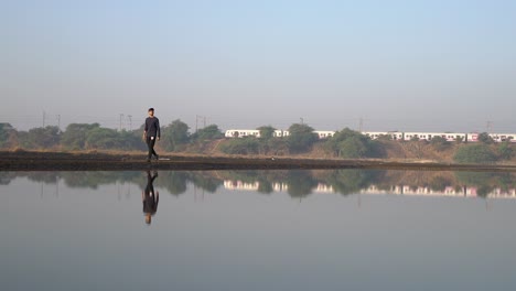 Hombre-Caminando-Por-El-Sendero-Junto-Al-Lago-Con-Reflejos-Sobre-El-Agua-En-Mumbai,-India