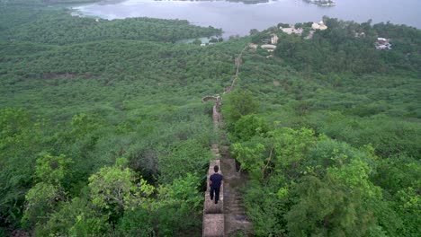 Male-Tourist-Overlooking-While-Standing-On-A-Concrete-Narrow-Path-In-Lush-Green-Valley-Leading-To-The-Beautiful-Lake-In-Udaipur,-Rajasthan,-India---Wide-Shot