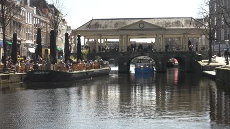 Boat-Passing-Under-Koornbrug-Bridge-Over-Rhine-River-With-People-Enjoying-Sitting-Outside-Beside-Cafe-In-Lieden