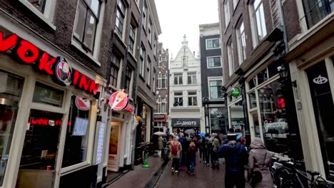 A-POV-shot-walking-down-the-narrow-crowded-Warmoesstraat-surrounded-by-restaurants-and-retail-shores-on-a-cold-rainy-day-in-Amsterdam,-Netherlands