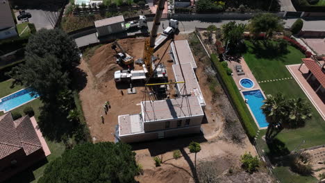 Rising-aerial-view-looking-down-at-crane-lifting-modular-housing-onto-building-site-foundation-plot