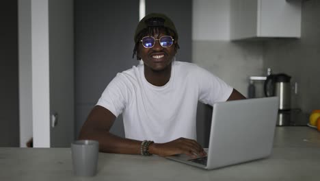 Young-black-male-vlogger-sitting-with-laptop-at-the-kitchen,-speaking-on-camera-while-livestreaming