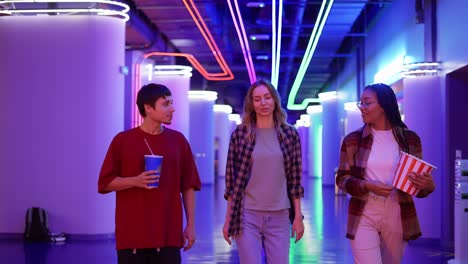 Group-of-three-friends-walking-by-cinema-hall-with-neon-interior-with-drink-and-popcorn-bucket