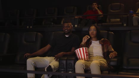 Mixed-raced-young-couple-sitting-in-movie-theatre-watching-film-and-taking-popcorn