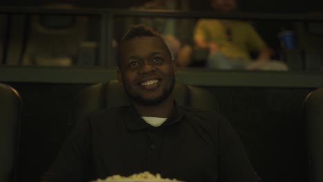 African-American-man-sitting-in-armchair-watching-a-comedy-at-the-cinema,-taking-popcorn
