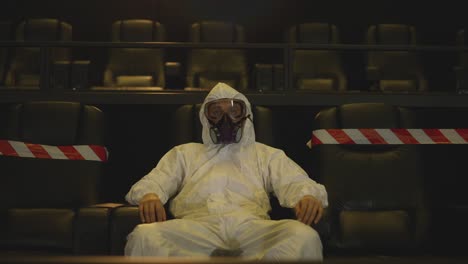 Portrait-of-a-man-in-white-protection-costume-and-respirator-sitting-alone-at-the-cinema