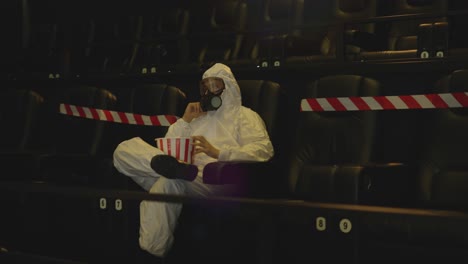 Portrait-of-a-man-in-white-protection-costume-and-respirator-sitting-alone-at-the-cinema-taking-popcorn
