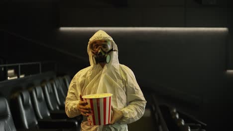 A-man-in-white-protection-costume-and-respirator-came-alone-to-the-cinema-with-bucket-of-popcorn,-slow-motion