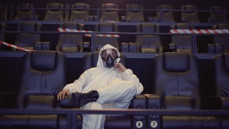 Portrait-of-a-man-in-white-protection-costume-and-respirator-sitting-alone-at-the-cinema,-front-view