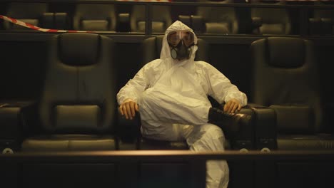 Relaxed-man-in-white-protection-costume-and-respirator-sitting-alone-at-the-cinema