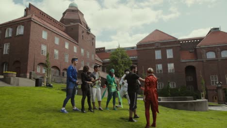 College-students-in-superhero-costumes-stand-and-play-on-a-grassy-hillside-near-large-brick-buildings-on-their-university-campus