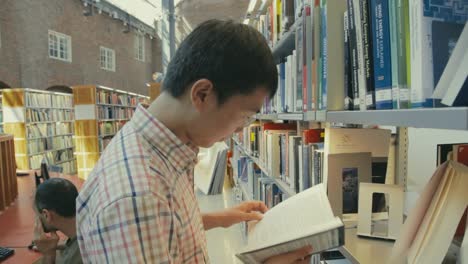 Un-Joven-Asiático-Mira-Un-Libro-En-El-Estante-De-La-Biblioteca-De-La-Universidad